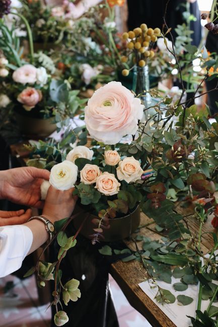 A florist's hand, wearing a white shirt and a silver watch, arranging a bouquet of delicate pale pink and cream ranunculus roses with lush green leaves and eucalyptus stems. The floral arrangement is