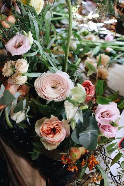 A detailed floral arrangement featuring a variety of fresh flowers including large, soft pink and peach ranunculuses, cream and blush lisianthus, and small orange accents, all complemented by lush gre