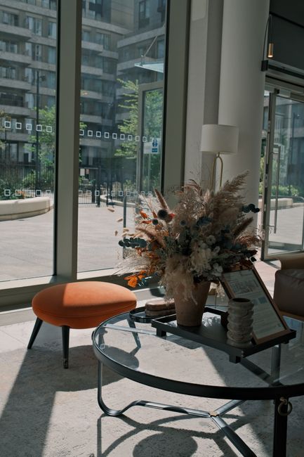 A large, lush floral arrangement featuring a mix of dried and preserved flowers, including pampas grass, muted-colored roses, and other delicate blooms in soft earthy tones, arranged in a tall, beige