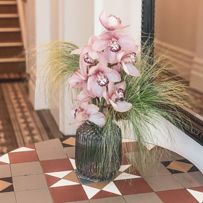 A tall floral arrangement featuring soft pink orchids with large, open blooms and white markings on the petals, placed in a dark, ribbed glass vase. The orchids are complemented by wispy green ornamen