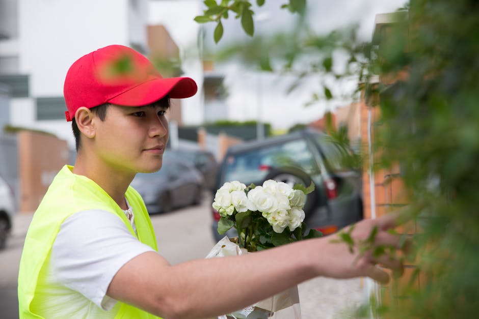 A male florist wearing a red cap and a high-visibility yellow vest arranges a bouquet of fresh white flowers, including roses and hydrangeas, wrapped in white paper with a ribbon. He stands outdoors b