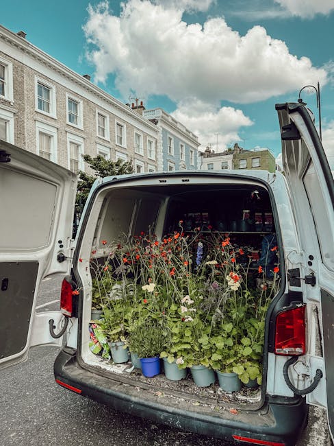 Open rear of a white delivery van filled with potted plants and blooming flowers, including red poppies and white daisies, arranged in light blue and grey pots. The van is parked on a street lined wit