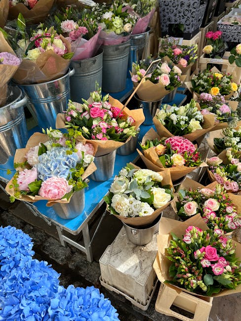 A collection of floral bouquets arranged in metal and white ceramic vases, featuring a variety of fresh flowers in pastel pinks, whites, and blues. The bouquets include roses, hydrangeas, and other bl