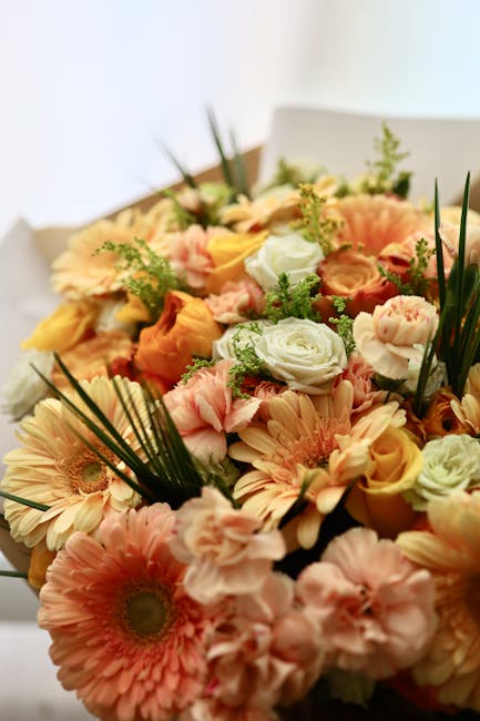 A vibrant floral bouquet featuring a mix of soft peach and cream-colored gerbera daisies, pale pink lisianthus, and white roses, accented with sprigs of greenery such as fern and long, slender blades 