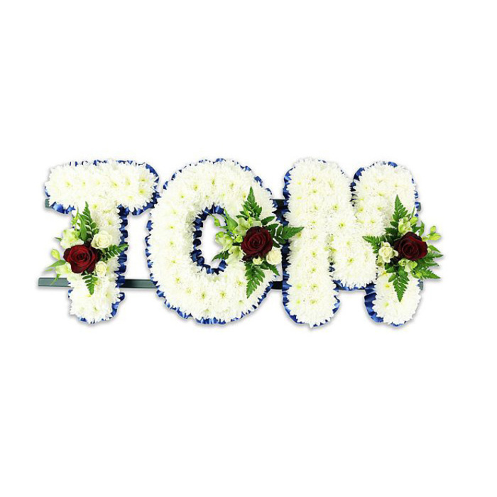 A carefully composed memorial display that spells TOM in sculptural, voluminous letters made entirely of small, pristine white chrysanthemum blooms. Each flower is positioned to form a dense, textured surface that reads soft and substantial; the overall silhouette is clean and formal. A striking royal blue ribbon outlines every letter, providing a smart, contrasting border that sharpens each edge and gives the tribute a refined visual punctuation against the stark white backdrop. Within the middle and lower portions of each letter, symmetrical accent sprays feature a single, richly hued red rose as the emotional centre, supported by clusters of creamy white spray roses and sprigs of fern-like foliage that add depth and a natural rhythm to the flat plane of chrysanthemums. The arrangement is mounted on a discreet dark metal framework running along the base, visible enough to suggest its purpose but finished to remain unobtrusive during display. The lighting is even and neutral, allowing textures and colour to be read without distraction - the chrysanthemum whites appear cool and comforting, the royal blue ribbon keeps a respectful crispness, and the red roses give an austere, heartfelt counterpoint. As a florist who has placed memorials for families near Nine Elms and the New Covent Garden Market, I recognise this as a classic, personalised tribute for funeral services and graveside commemorations: carefully handcrafted, reverent in tone, and intended to offer both a visual sign of remembrance and a tactile expression of shared sorrow.