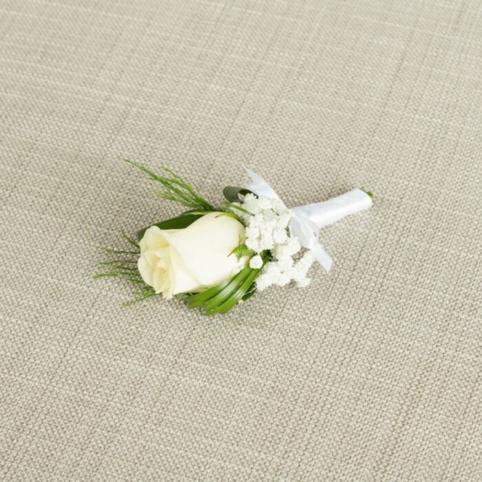 A classic wedding boutonniere lies diagonally across a pale beige fabric that looks like natural linen, its subtle woven pattern providing a warm, understated backdrop similar to the stylish interiors of modern Nine Elms venues. At the centre of the composition is a single white rosebud in a creamy ivory tone, its petals tightly coiled yet edged with a hint of opening, suggesting a bloom chosen at just the right moment of freshness. The rose's surface appears velvety and smooth, catching the soft, diffused light so that each curve and fold is gently defined without harsh shadows. Surrounding the rose on one side, clusters of fine white baby's breath create a cloud-like fringe, their miniature blossoms adding intricate texture and a sense of airiness, like a soft mist of flowers. Threaded among the rose and gypsophila are slender, rich green leaves and a few fern-like sprigs, some glossy and linear, others feathery and more delicate, giving the boutonniere natural depth and a quietly lush feel. The stems are carefully aligned and tightly wrapped in a band of pure white satin ribbon, the fabric smooth and slightly reflective, finishing in a small, tidy bow placed just beneath the flower head. This crisp bow indicates the piece is ready to be pinned neatly onto a suit lapel for a ceremony along the Thames or a reception near the new Nine Elms developments. The lighting remains soft and even across the scene, enhancing the contrast between the creamy white petals, the cloud of baby's breath, and the vibrant green foliage while keeping the background gently blurred. The overall effect is one of polished simplicity and timeless romance, perfect for illustrating groom's accessories, bridal party styling, or refined boutonniere design on wedding planning pages.