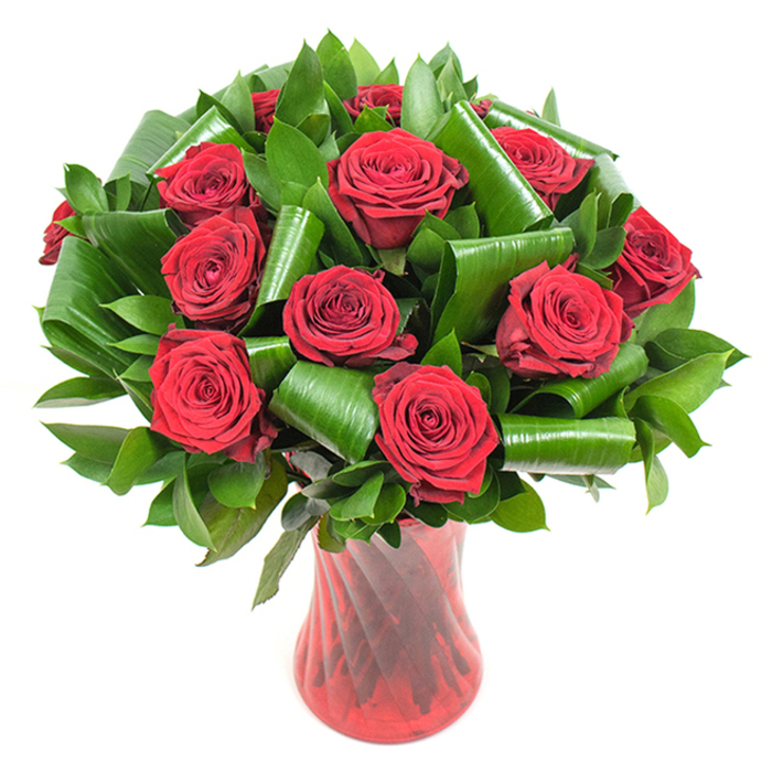 Framed against a crisp white backdrop, this carefully constructed bouquet features a generous cluster of deep crimson roses-approximately a dozen or more-each bloom in full, velvety bloom with inward-spiralled petals that form dense, tactile cups. The roses are nestled within abundant dark-green foliage: a dense lower layer of glossy Salal- or Ruscus-type leaves provides solidity and a dark, even contrast to the flowers, while larger, bright green Aspidistra-style leaves have been folded and rolled into polished scrolls and tubular coils. These sculpted greens act like living architecture, cradling the rose heads and creating a pleasing dialogue between soft petals and crisp leaf edges. The stems rest in a tall, translucent ruby-red glass vase whose subtle twisted or ribbed surface throws vertical highlights and mirrors the bouquet's passionate palette; the vase tapers slightly toward an elegant waist and flares gently at the mouth, giving the arrangement stable support and a graceful silhouette. Soft, even lighting accentuates texture-the velvet of the petals, the glossy sheen of the leaves, and the cool clarity of the glass-while the white setting isolates the composition so every nuance reads clearly. As a practising florist in Nine Elms and the surrounding Battersea and Vauxhall neighbourhoods, I recognise this as a classic romantic piece, ideal for anniversaries, Valentine's messages or any occasion calling for a confident, timeless expression of love. Flower Delivery Nine Elms prepares stems like these with attention to balance, scent and long-lasting freshness.