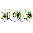 The photograph presents a bespoke TOM funeral tribute, three large floral letters displayed on a narrow dark base so they appear to stand in a neat row against a plain white background. Each capital letter is densely filled with snowy white chrysanthemums, the blooms pressed close together to create a smooth, cushion-like surface that looks soft yet formally arranged. The outer contours of the T, O, and M are edged with a royal blue ribbon, its satin folds forming a crisp, continuous border that clearly defines the lettering and introduces a calm, dignified contrast to the white flowers. To bring warmth and individuality, the florist has added small accent sprays to each letter: clusters of velvety deep red roses, paired with tiny white or cream spray roses and slender, trumpet-shaped white flowers similar to freesia, all surrounded by mixed greenery. Delicate fern fronds and glossy leaves extend around these clusters at the top-left of the T, along the curve of the O, and near the upper-right section of the M, creating focal points that guide the eye across the tribute. The design feels perfect for a funeral or memorial service in Nine Elms, perhaps set near the front of a modern chapel or community space overlooking the river. One can picture the tribute resting quietly beside candles and photographs, the faint scent of fresh flowers rising from the chrysanthemums and roses. This TOM arrangement speaks of affection and respect, its balance of white, blue, and red echoing themes of purity, serenity, and enduring love within the evolving urban landscape of Nine Elms.