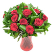 A lush, rounded bouquet of twelve to fifteen deep crimson roses sits as the image's quiet hero, each bloom opened to show velvety, spiralled petals that catch the light with a soft, satiny sheen. The rich, wine-red tones of the roses are intensified by even studio lighting, which reveals subtle gradations from near-maroon at the petal bases to a brighter crimson at the edges. Around and beneath the roses is an abundance of dark green foliage: dense, glossy leaves that form a verdant backdrop and smaller, leathery Ruscus- or Salal-like fronds that give the cluster weight and fullness. Larger, emerald Aspidistra-style leaves have been skillfully rolled and looped into architectural cylinders and soft spirals, their smooth surfaces reflecting narrow highlights and adding a contemporary flourish to the classic red rose arrangement. The stems sit in a tall, slender, translucent ruby-red glass vase with a faint ribbed twist, vertical lines running the length of the vessel so it gleams where the light strikes, echoing the deep red of the blooms while remaining delicate and refined. Set against a clean white background, the bouquet's colour and form are isolated and amplified, making the reds and greens feel almost tactile; one imagines the velvety petal texture, the cool glass, and the gentle perfume of fresh roses. The composition reads as a celebration of romantic tradition with modern styling-an ideal image for talk of anniversaries, intimate gifts, and elegant centrepieces-as if prepared by a florist who knows how to let colour and structure speak together.