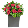 In this elegantly modern composition, a cluster of rich, velvety red roses forms the immediate focal point, each bloom opening in generous spirals that read like soft red velvet against the eye. The roses are nestled among broad, glossy green leaves that have been carefully curled and intertwined to create a rhythmic, sculptural backdrop; the foliage's waxy sheen catches the bright, clear studio light so that edges glimmer and shadows fall with gentle clarity. The arrangement sits in a crisp, matte black cube whose geometric lines ground the display and offer a contemporary counterpoint to the organic curves of petals and leaves. Lighting here is steady and clean, lifting the deep crimson into almost jewel-like tones while revealing subtle texture in each petal and the cool clarity of the leaves.There is an intimate sensory suggestion in this bouquet: a soft, classic rose fragrance balanced by fresh, almost green notes from the foliage, creating an air that is both romantic and refreshingly clean. As a florist familiar with Nine Elms and the riverside developments around Battersea Power Station and Embassy Gardens, I picture this piece set on a sleek console in a modern apartment or brought as a considered anniversary gift after dinner along the Thames. The design reads as both a declaration and an elegant accent-ideal for marking romantic milestones, offering gratitude, or brightening a neat urban interior-each detail intentionally composed to make a quiet, memorable impression on sight and scent.